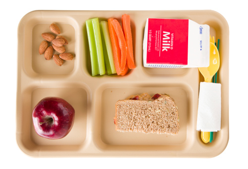 An overhead close up of a tray containing a healthy school lunch. It consists of crunchy peanut butter and jelly sandwich on whole wheat multigrain bread, celery sticks, carrot sticks, almonds, a red apple and milk. Isolated on white.